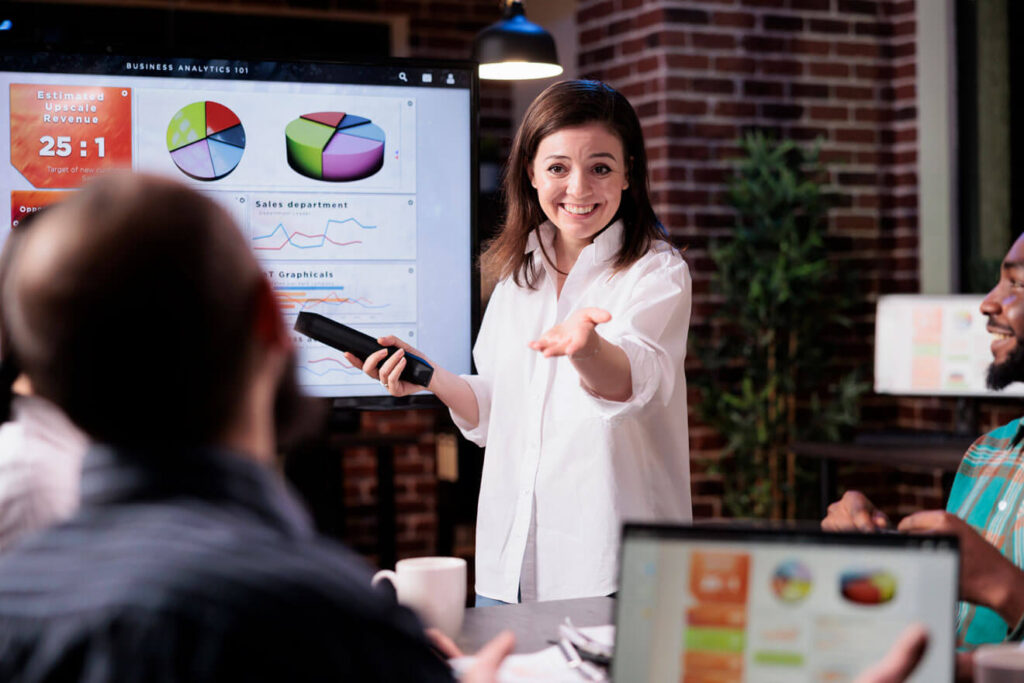 mulher executiva de vendas sorrindo em frente ao monitor apresentando seu pitch de vendas para um homem e segurando um laptop que exibe pie charts e gráficos analíticos